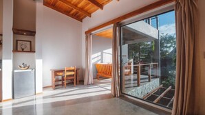 Um quarto espaçoso com uma grande porta de vidro deslizante que dá para uma varanda. Há uma mesa e cadeiras de madeira, um refrigerador pequeno e um quadro pendurado na parede.