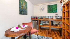 A compact kitchen with wooden cabinets, a dining table with a chair, and two wall-mounted fans. - The Surf Shack - With direct beach access (Playa Guiones)