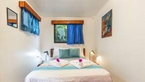 A bedroom with a wooden bed frame, a window with blue curtains, a bedside lamp, and a painting on the wall. - The Surf Shack - With direct beach access (Playa Guiones)