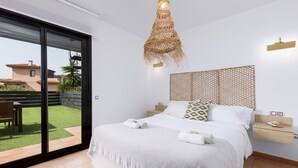 A bedroom with a large bed, a hanging light fixture, a small bedside table, and a view of a green outdoor area. - Home2Book Casa Calma Garden, Pool & Relax (Majanicho)