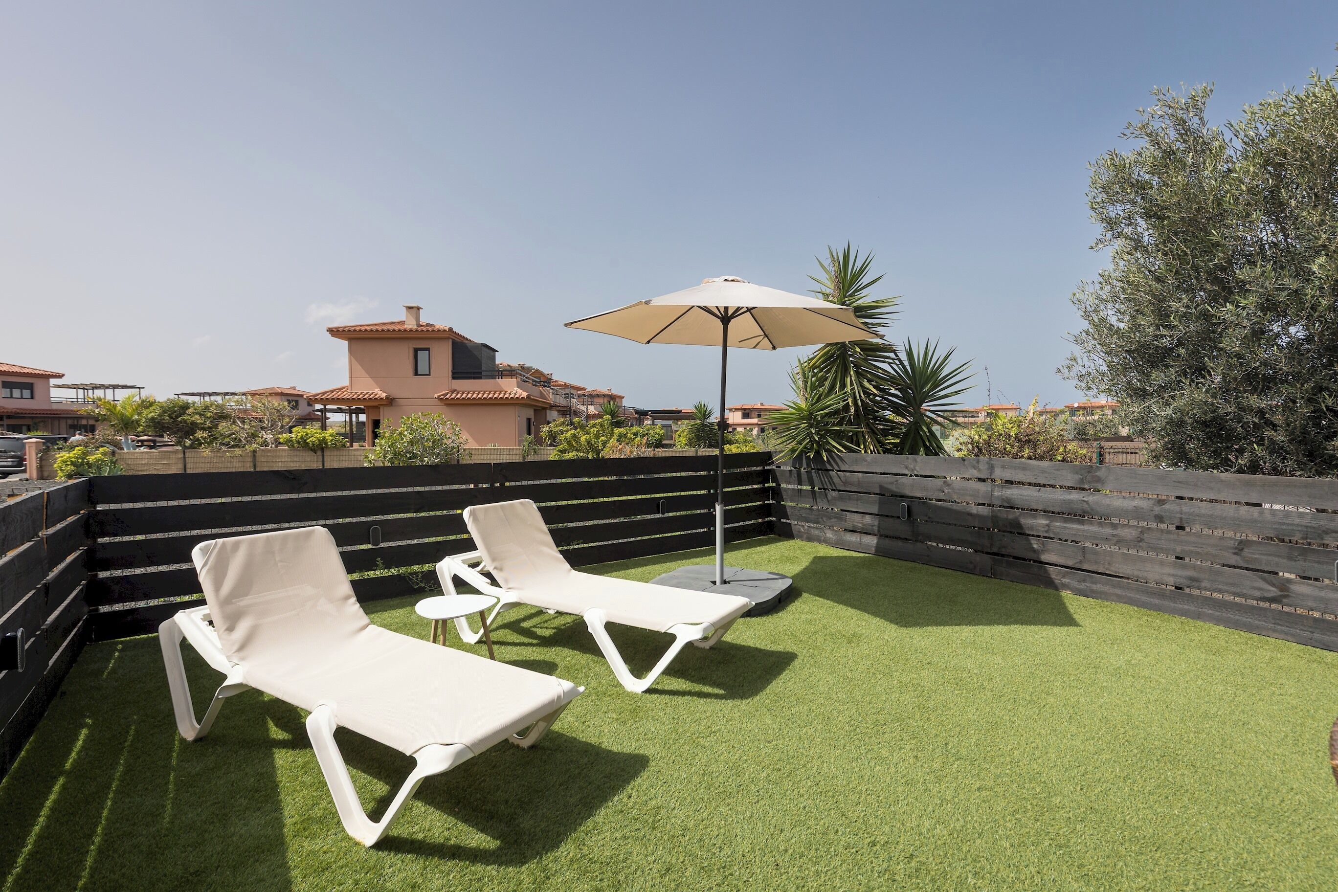 Eine Dachterrasse mit künstlichem Rasen, zwei weißen Liegestühlen, einem Sonnenschirm und einem Holenzaun.