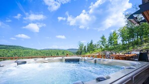 A hot tub with a wooden deck and outdoor seating, surrounded by a forested landscape. - Charming log cabin nestled in the woods, with a spa and in-ground pool.  (Mandeville)