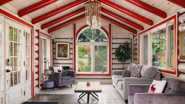 A cozy living room with a high ceiling, wooden beams, and large windows offering a view of greenery outside. - Charming log cabin nestled in the woods, with a spa and in-ground pool.  (Mandeville)