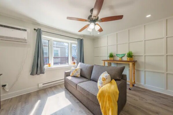 Een woonkamer met een grijze bank, een plafondventilator, een raam met gordijnen en een klein houten tafeltje met een plant.