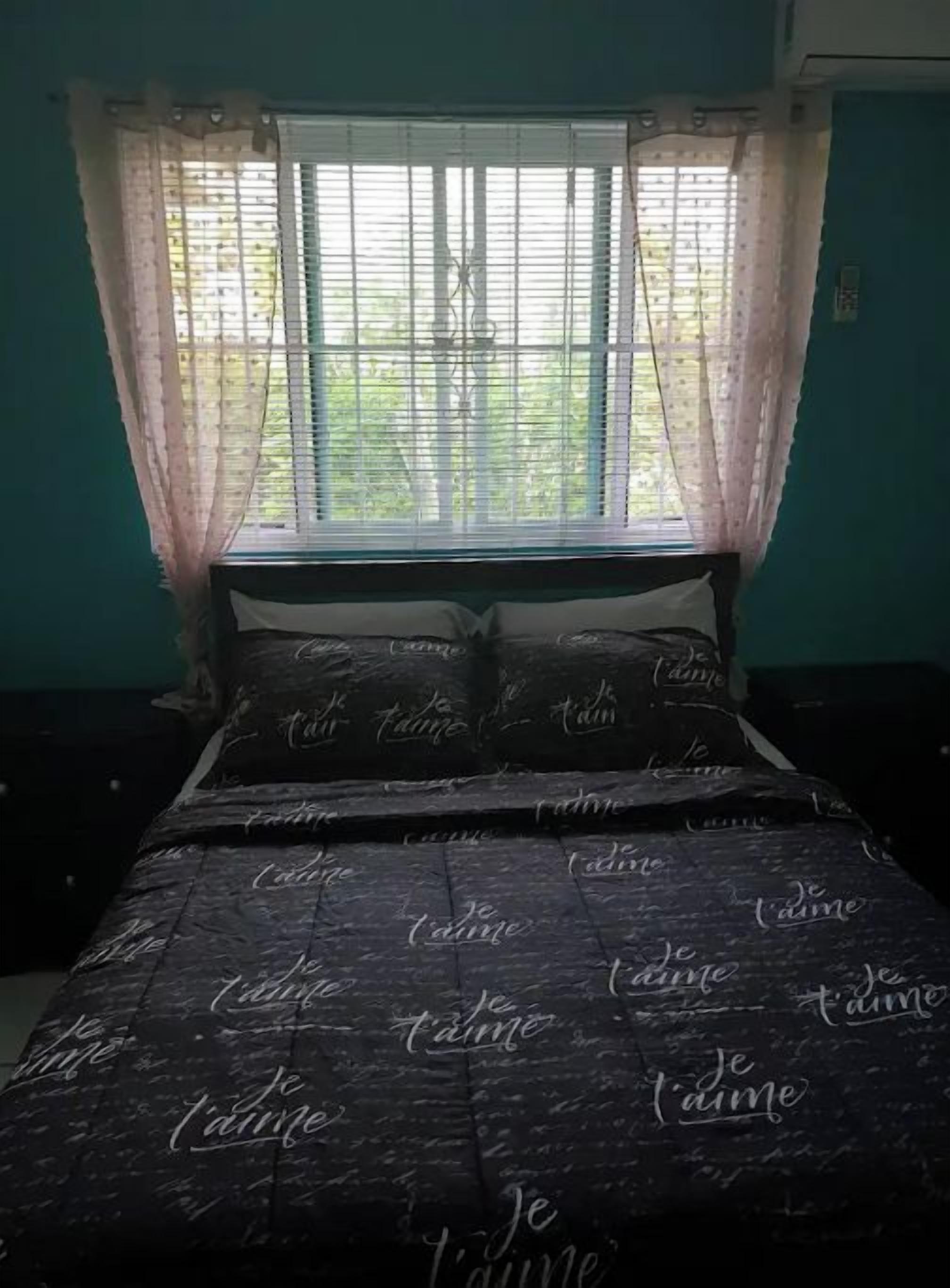 A bedroom with a bed covered in a dark sheet with 'Je t'aime' printed on it, a window with sheer curtains, and teal walls.