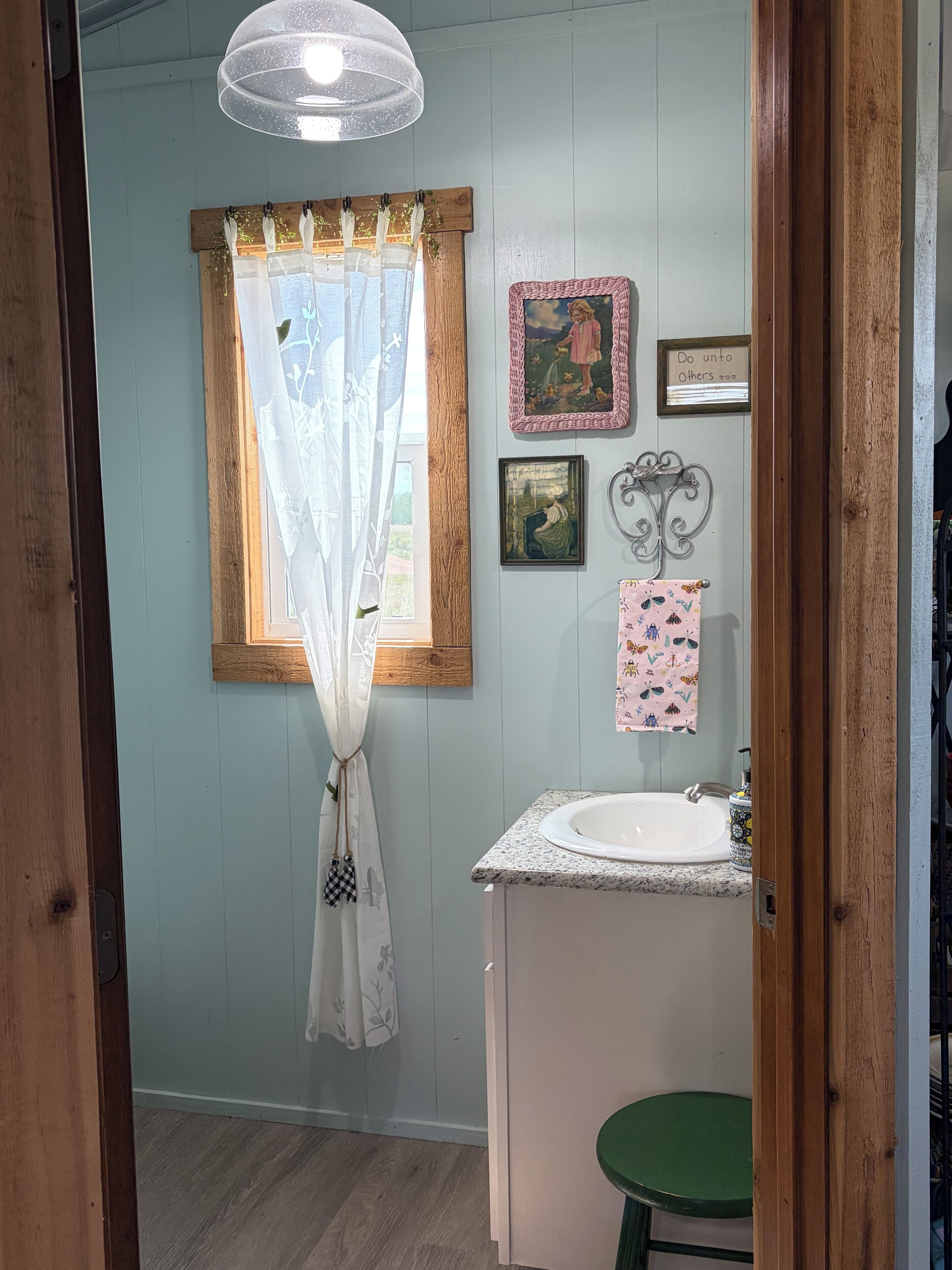 Een badkamer met een wastafel, een groene kruk, een raam met een dunne gordijn en een muur met ingelijste foto's en een bord.