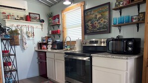 A kitchen with white cabinets, stainless steel appliances, and a microwave. There are shelves with various items and a window with blinds.