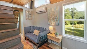 Une pièce chaleureuse avec un escalier en bois, un canapé gris, une petite table avec une lampe et une grande fenêtre donnant sur un paysage verdoyant.