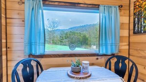 A wooden cabin room with a table, two chairs, a window with curtains, a small plant, and a candle.
