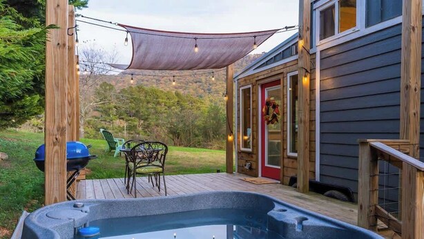 A hot tub on a wooden deck with a covered patio, a barbecue grill, and a dining area with chairs.