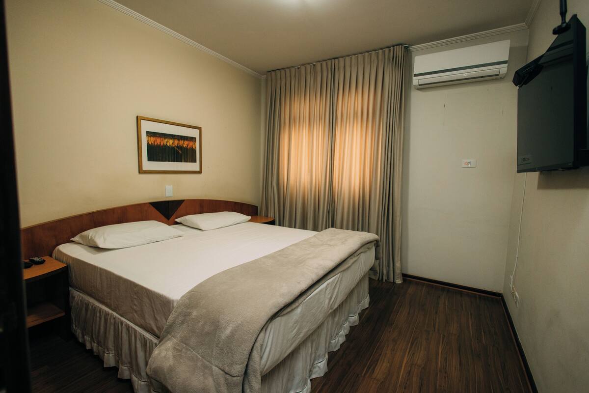 A hotel room with a bed, a nightstand, a TV, and an air conditioning unit.