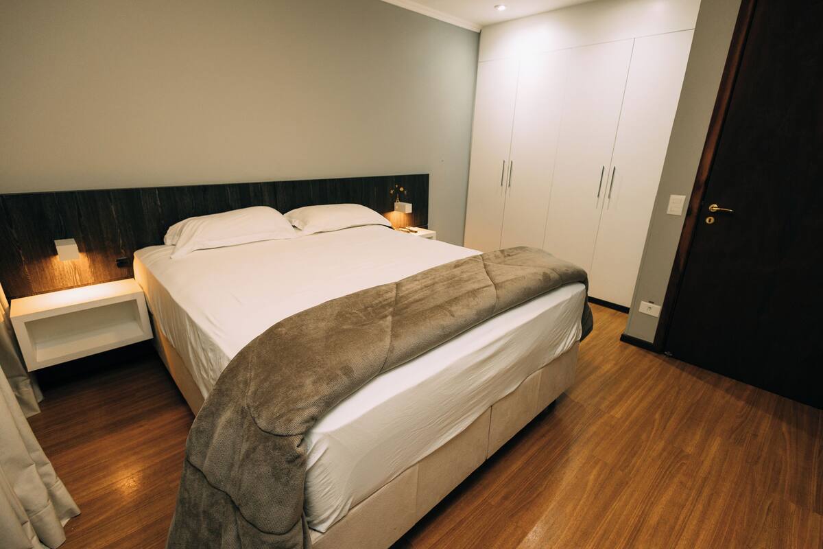 A modern bedroom with a large bed, wooden flooring, a nightstand, and a wardrobe.