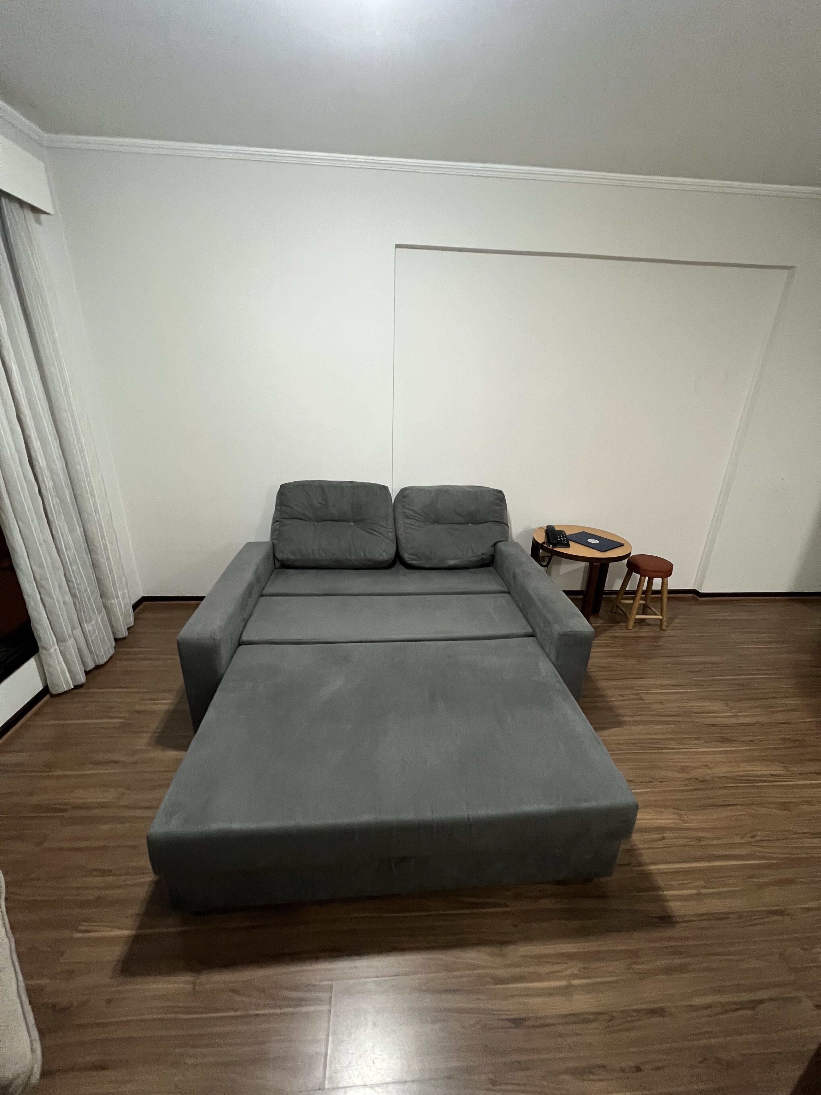 A grey sofa bed with two pillows, a small wooden side table with a remote control and a book, and a wooden floor.