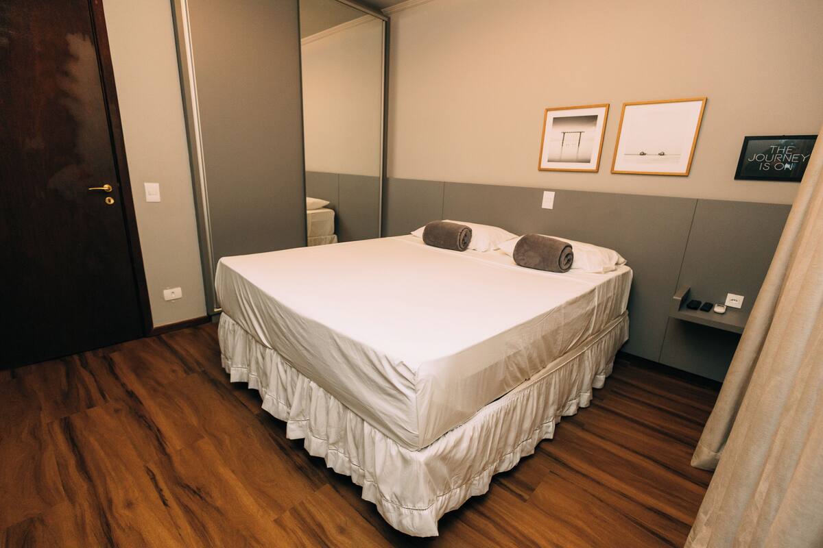 A neatly made bed with a white bedspread and ruffled skirt, a wooden floor, a door, and a wall with framed pictures.