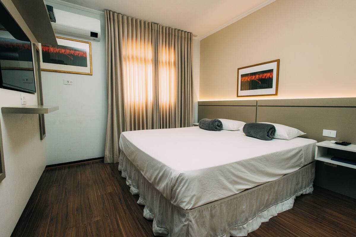 A hotel room with a large bed, a TV mounted on the wall, a bedside table, and a window with curtains.