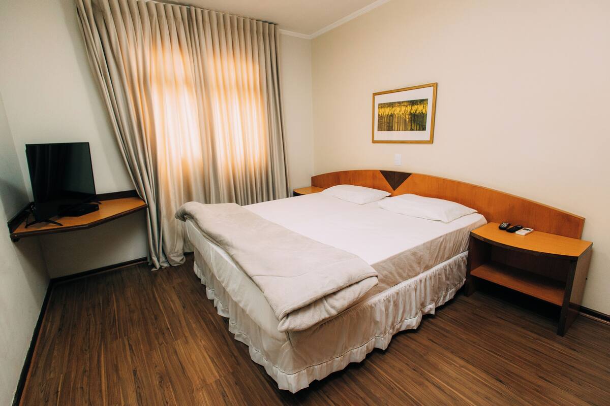 A hotel room with a bed, a desk with a television, a nightstand, and a painting on the wall.