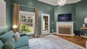 A living room with a teal sofa, a fireplace, a TV mounted above it, and a large window with curtains. - Beautiful 3-Bdrm Home <> Park 2 Cars <> King Bed (Alexandria)