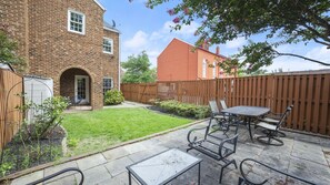 Un jardin avec une maison en briques, une clôture en bois, une terrasse avec une table et des chaises, et une pelouse.