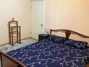 A bedroom with a bed, a wooden chair, a nightstand with a clock, and a door.