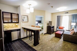 Ein kompaktes Hotelzimmer mit Küchenzeile, Essbereich, Wohnzimmer mit Sofa und Hocker sowie Blick durch eine Tür.