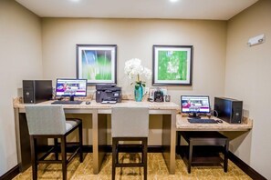 Een hotelkamer met een bureau, twee computers, een printer, een vaas met bloemen en ingelijste foto's aan de muur.