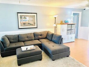 A living room with a sectional sofa, an ottoman, a framed picture on the wall, a lamp, and a cabinet with drawers. - Coastal Coconut Cove near Anna Maria Island (Bradenton)