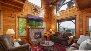 A cozy log cabin interior with a stone fireplace, leather furniture, and a scenic view through the open door. - Endless Views - King Beds - Close to attractions ! (Sevierville)