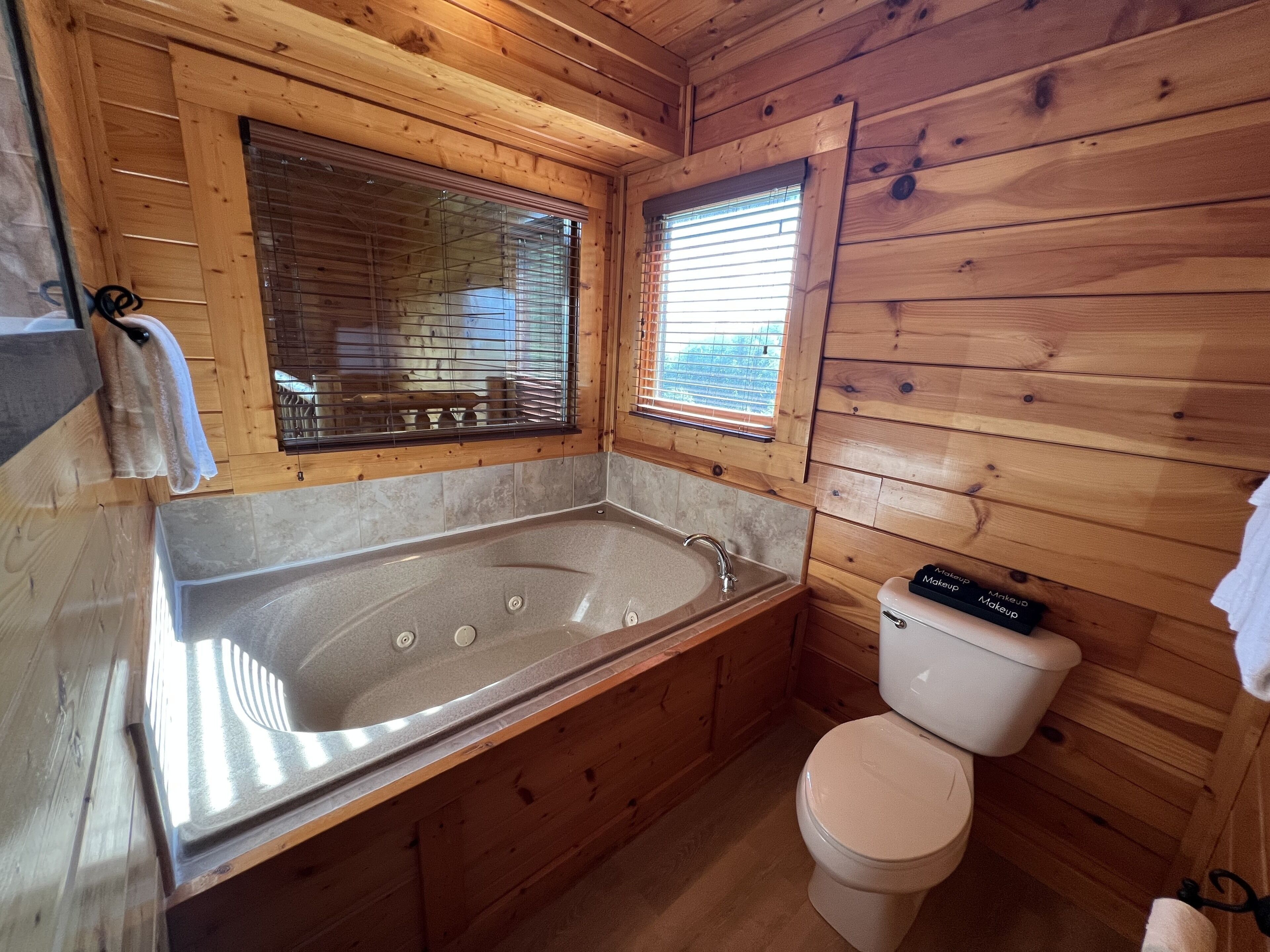 A wooden cabin bathroom with a bathtub, toilet, and a window with blinds.