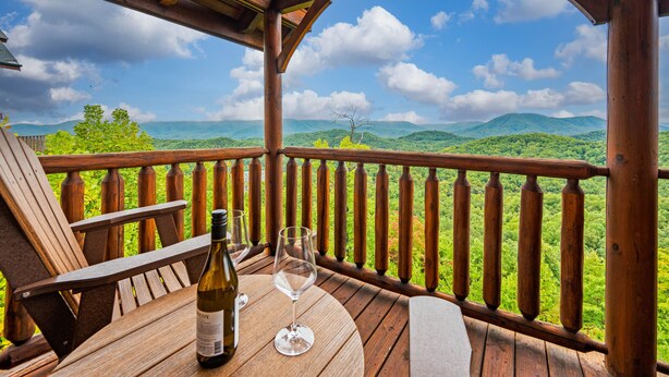 A wooden deck with a table, a bottle of wine, and a glass. The deck overlooks a mountainous landscape with lush greenery. - Endless Views - King Beds - Close to attractions ! (Sevierville)