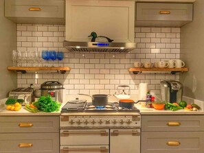Une cuisine équipée d’une cuisinière, d’un four et de divers appareils électroménagers. On y trouve des étagères avec de la verrerie et une statuette de canard accrochée au mur.