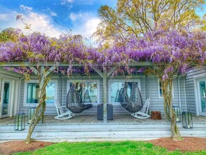 Un porche cubierto con flores moradas colgantes, dos sillones mecedores y una terraza de madera.