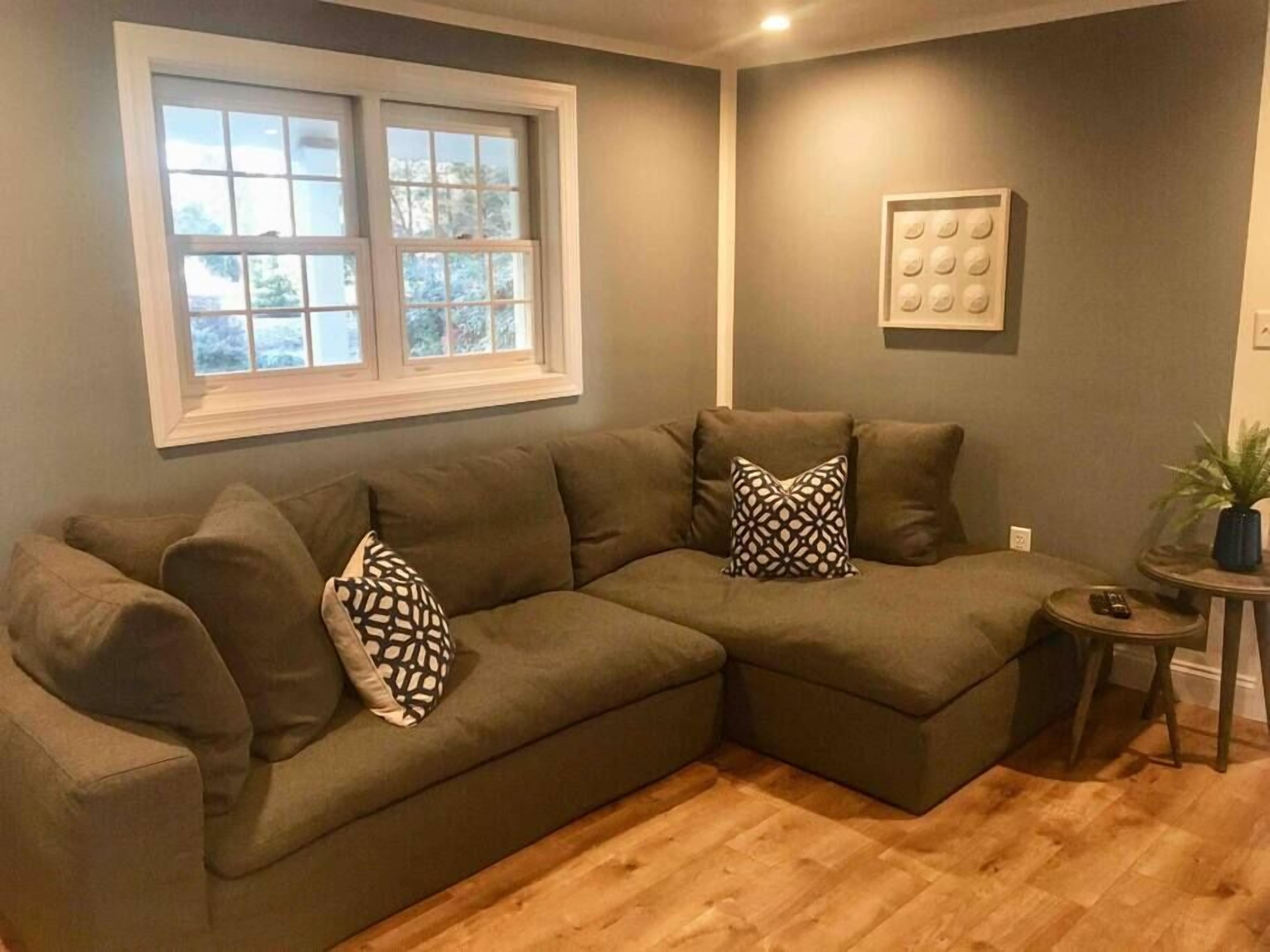 A sectional sofa with patterned cushions, a small side table, a potted plant, and a window with blinds.