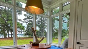 A room with a large window, a wooden table with a lamp, a bench with a cushion, and a basket with items. - Thorneton Point Estate (Easton)