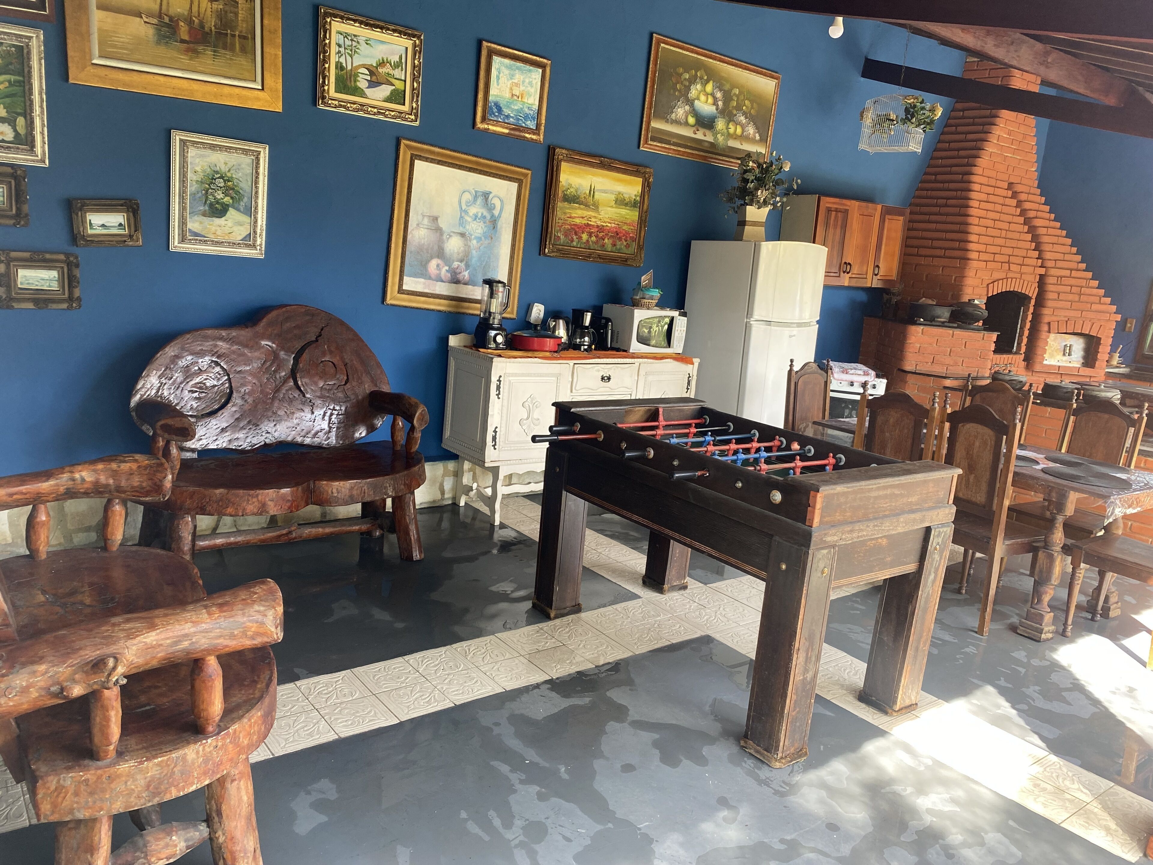 Een kamer met een tafelvoetbaltafel, houten stoelen, een bank, een keukenhoek met apparatuur, en diverse ingelijste foto's aan de muren.