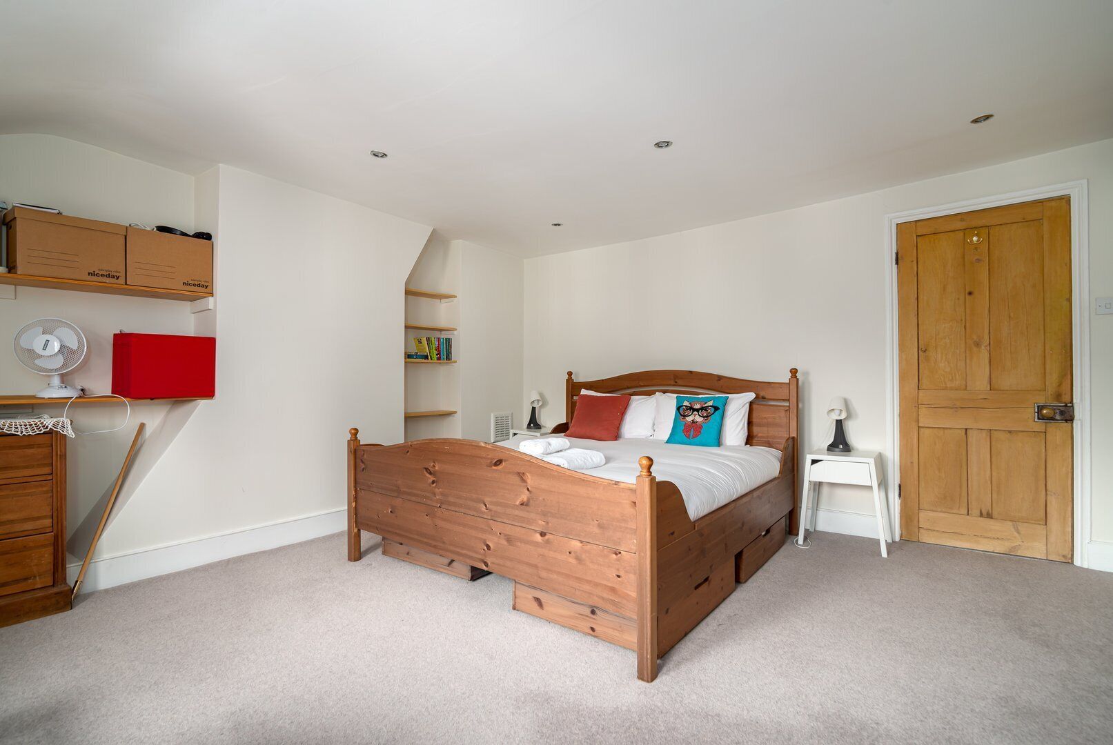 A bedroom with a wooden bed, a red cabinet, a white fan, and a wooden door.