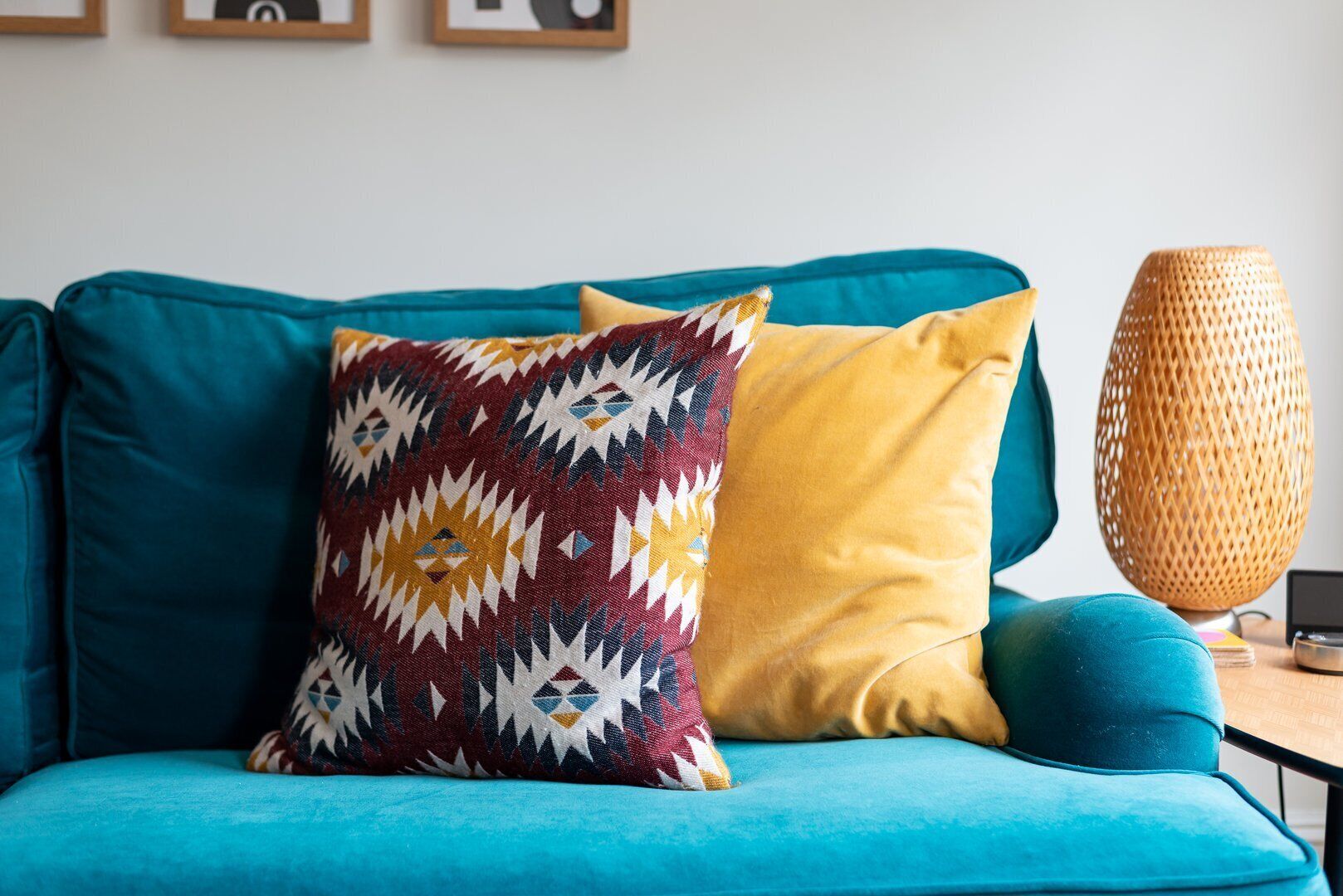 Un sofá color turquesa con cojines estampados y lisos, una lámpara trenzada y una mesita con un libro y un dispositivo.