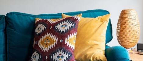 Un sofá color turquesa con cojines estampados y lisos, una lámpara trenzada y una mesita con un libro y un dispositivo.