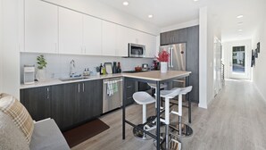A modern kitchen with white cabinets, a central island with bar stools, stainless steel appliances, and a view into the adjacent living area. - Cheerful 3-bedroom New Townhouse near UW/Seattle Children's, Free Parking, AC (Seattle)
