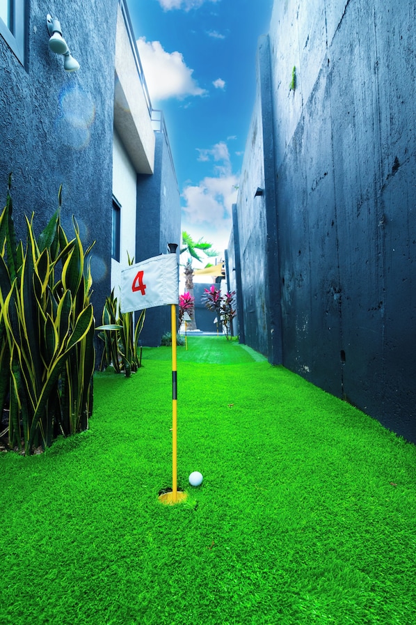 Een smalle steeg met een groen gazon, een golfgat gemarkeerd met een vlag, en een gebouw met een blauwe gevel.