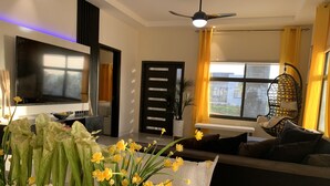 A modern living room with a flat-screen TV, a ceiling fan, a large window with curtains, and a decorative plant arrangement. - Ocho Rios Heights Ultra Lux Villa (Ocho Rios)