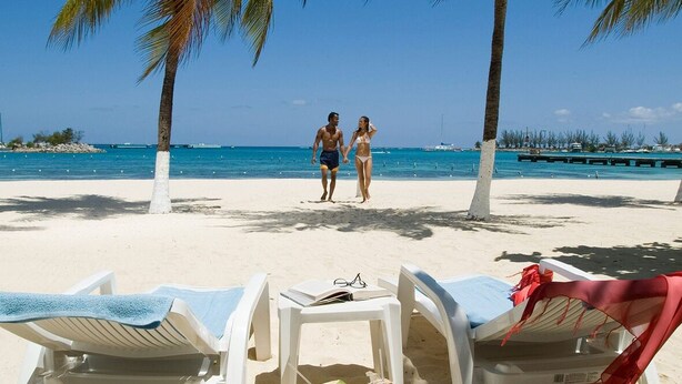 A beach with two people walking, palm trees, and lounge chairs with towels. - Ocho Rios Heights Ultra Lux Villa (Ocho Rios)