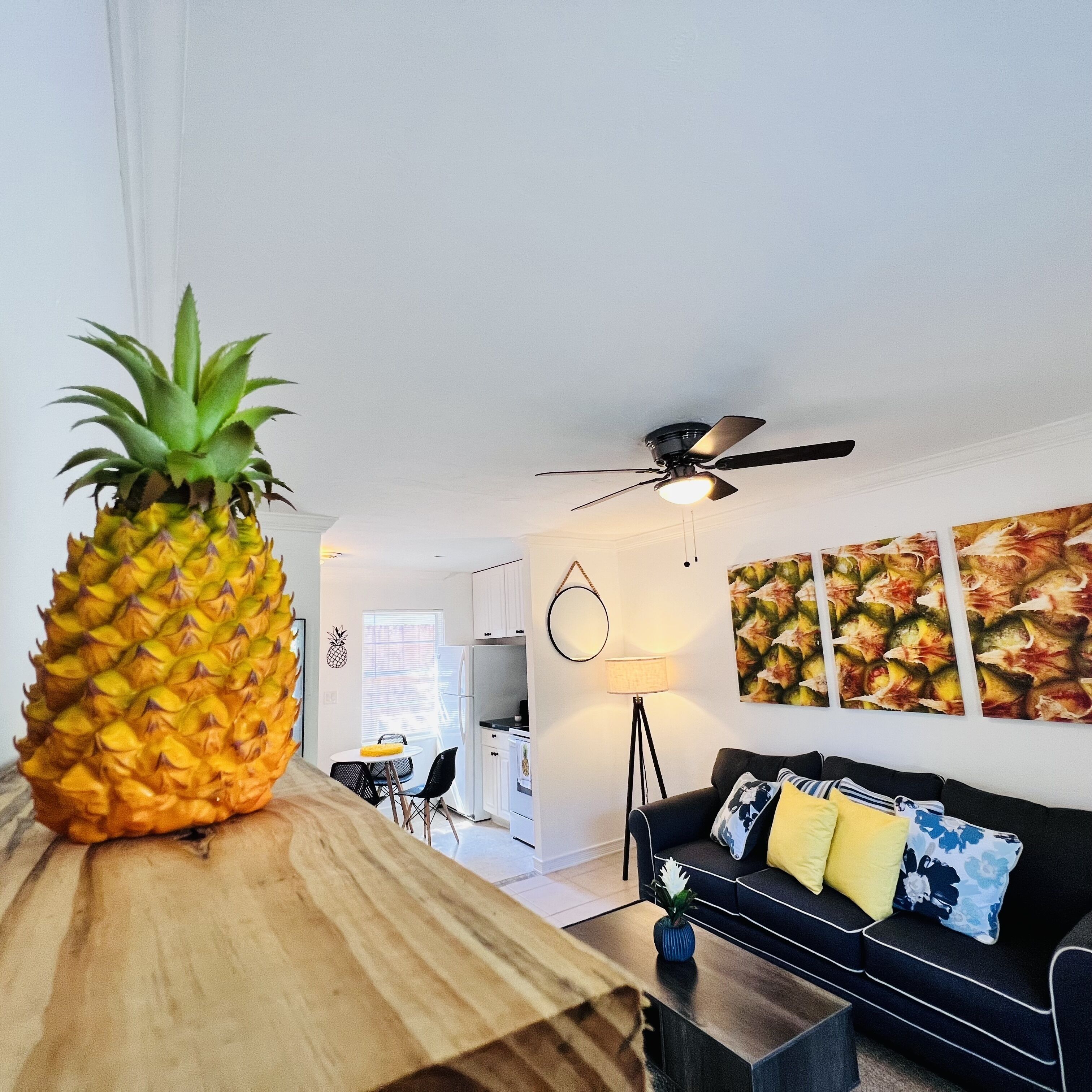 Ett vardagsrum med ett soffbord i trä, en svart soffa med gula och blå kuddar, en takfläkt och en ananas på bordet.