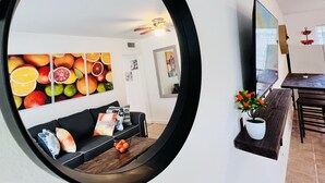 A modern living room with a large mirror, a sofa with cushions, a dining area with a table and chairs, and fruit wall art. - Coco Bay Condos (Fort Lauderdale)