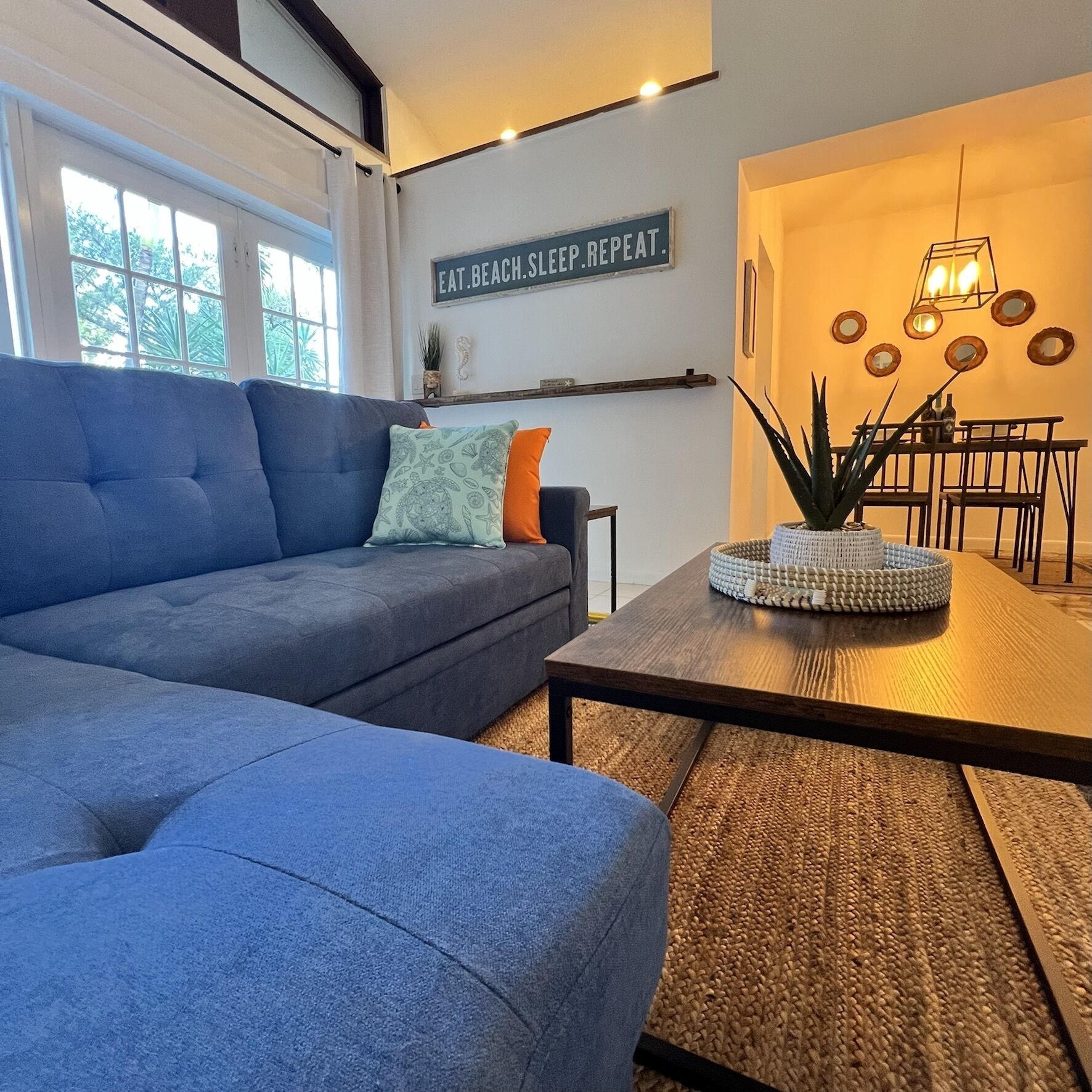 Un salon moderne avec un canapé bleu, une table basse et un panneau mural sur lequel on peut lire : « MANGER. SE PRÉLASSER À LA PLAGE. DORMIR. RECOMMENCER. »