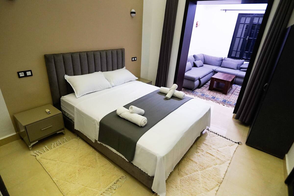 A neatly made bed with a grey headboard, a small bedside table, and a white bedside lamp. There is a view of a balcony with a grey sofa and a small wooden table.