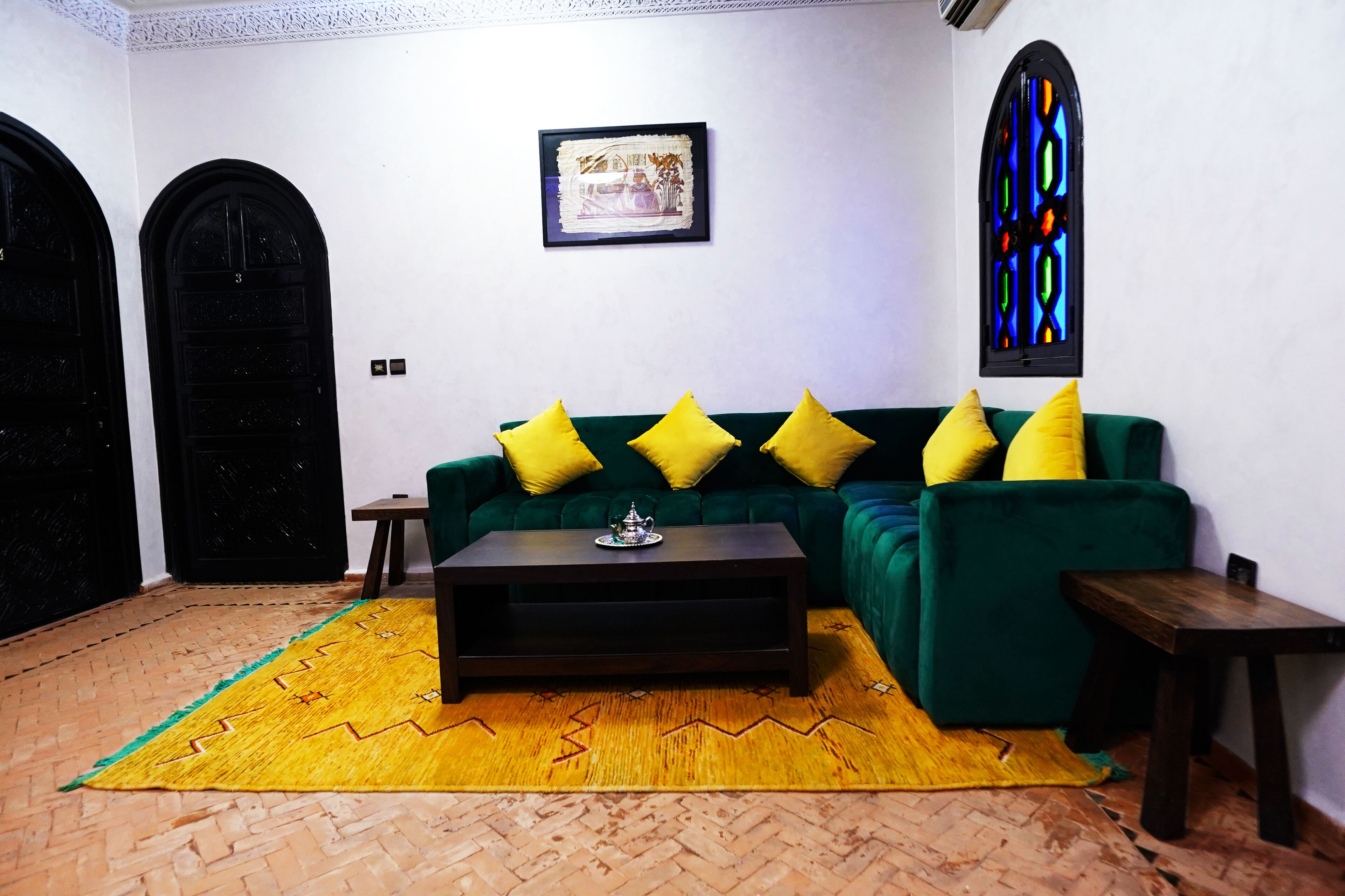Une pièce avec un canapé vert, des coussins jaunes, une table basse en bois, un tapis à motifs et une fenêtre à vitraux.