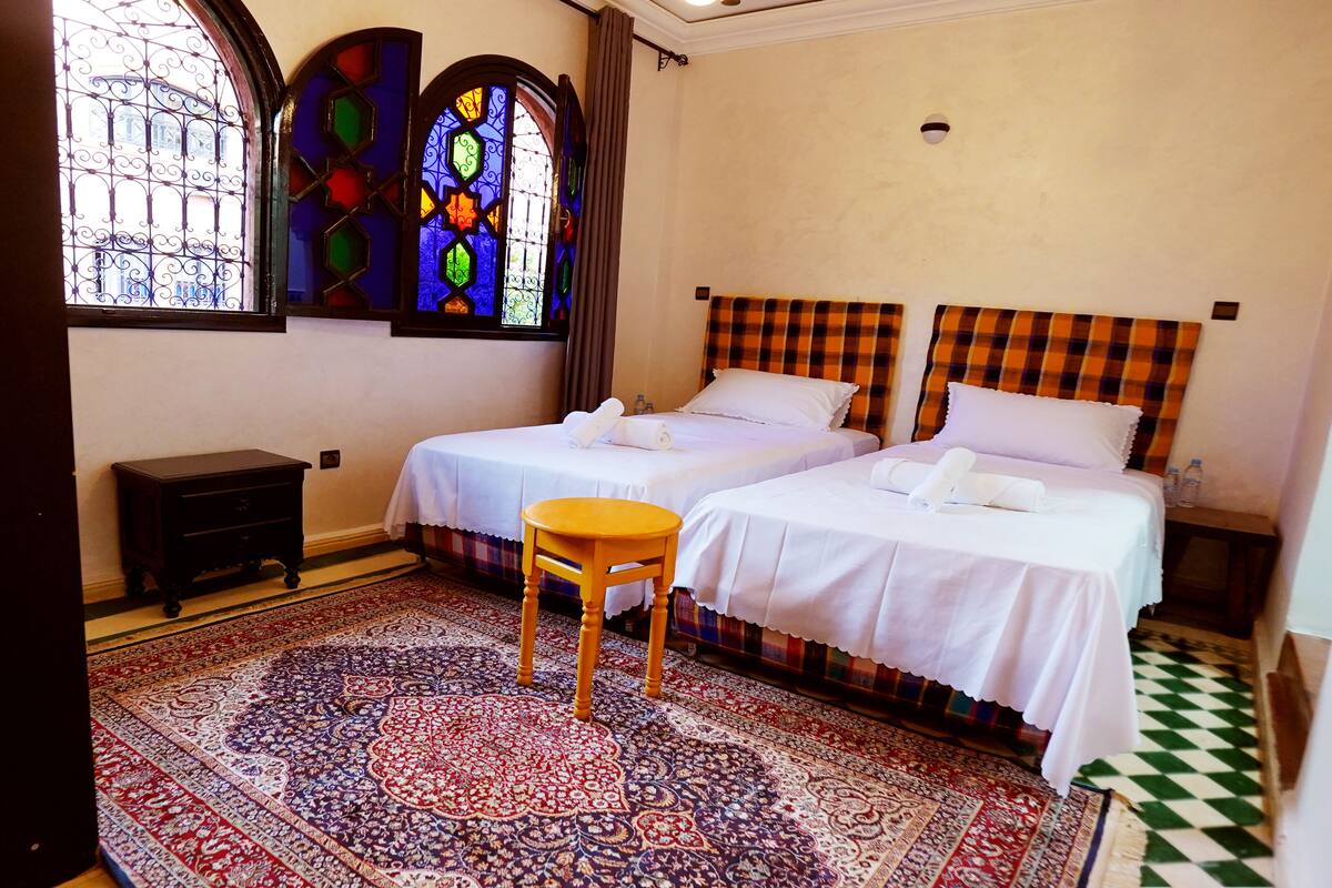A hotel room with two beds, a small wooden stool, a patterned rug, and stained glass windows.