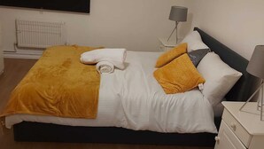 Un lit bien fait, recouvert d’une couette blanche et d’un plaid de couleur moutarde. On y trouve une petite table de chevet avec une lampe, ainsi qu’une commode de chevet dotée d’un tiroir.