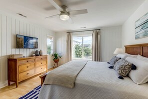 A bedroom with a bed, a wooden dresser, a television, a ceiling fan, and a window with curtains.