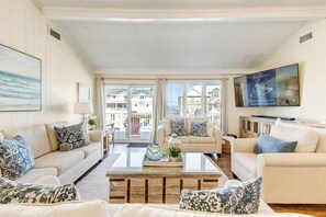 A spacious living room with two sofas, a coffee table, and a TV. The room has large windows with a view of other buildings. - Ohana | 350 ft to the beach | Private Pool, Hot Tub (Virginia Beach)